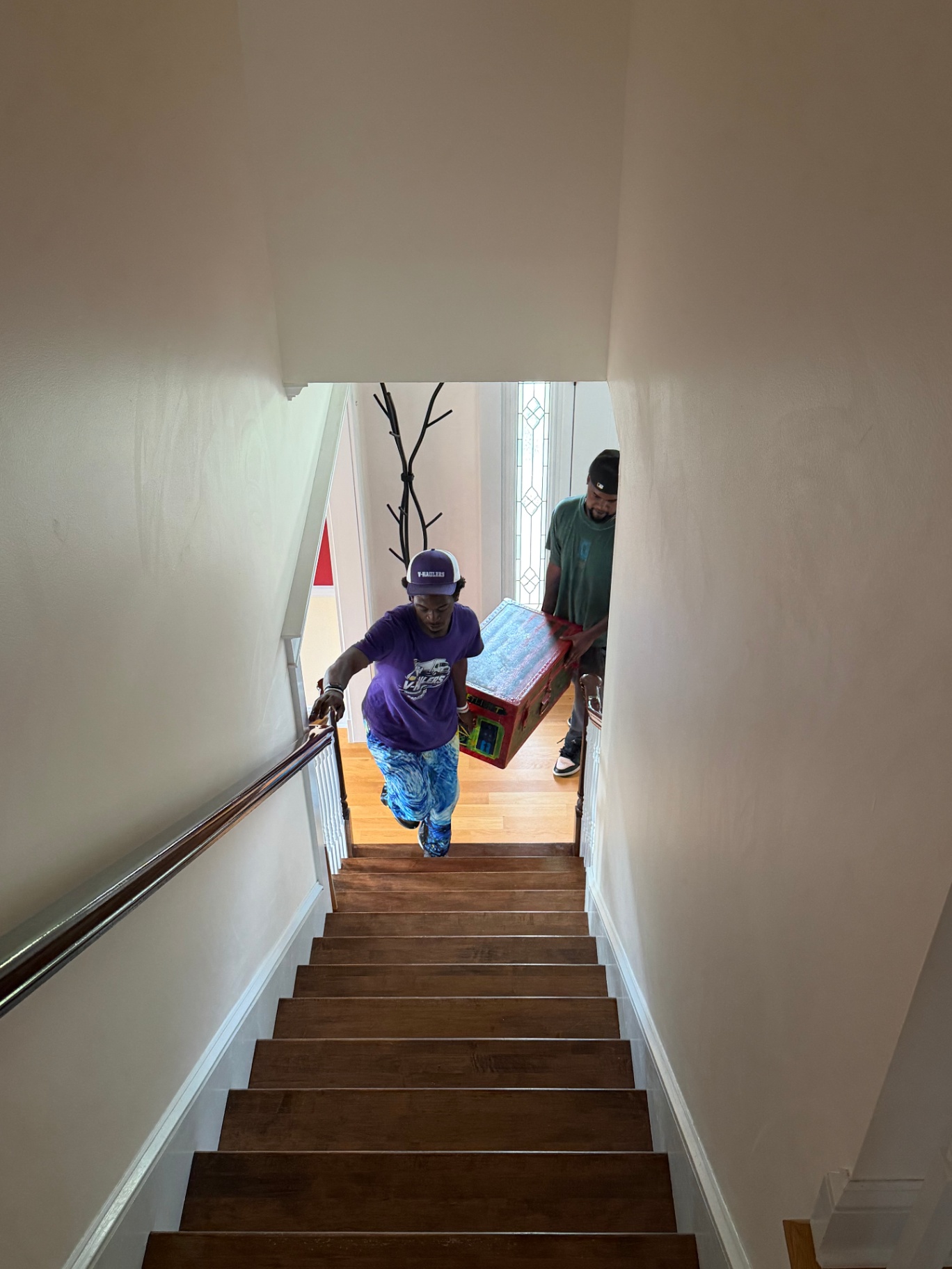 Two V-Haulers movers in purple shirts carrying a trunk up a staircase